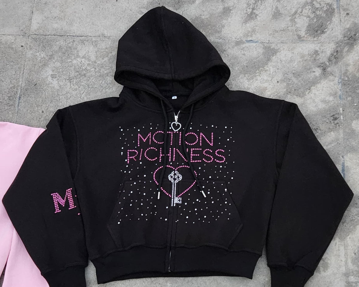 A black cropped zip-up hoodie with 'ACTION PLEASURE' and a key graphic in pink and silver rhinestones on the front, laid flat on a gray concrete floor next to a pink garment.